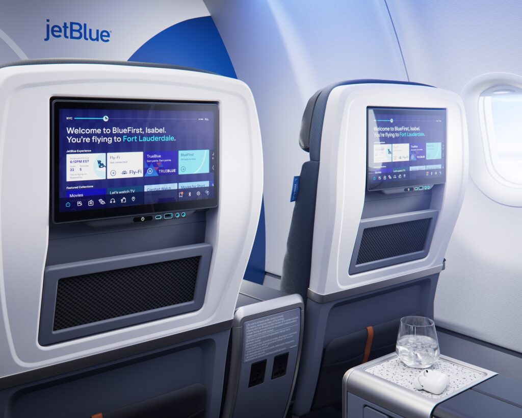 Two BlueFirst seatback entertainment screens displaying JetBlue’s inflight interface, with power outlets and a side table visible.