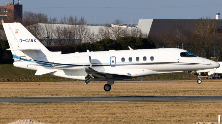 A white private jet with blue stripes and German registration D-CAWK lands on a runway, with industrial buildings and trees in the background.