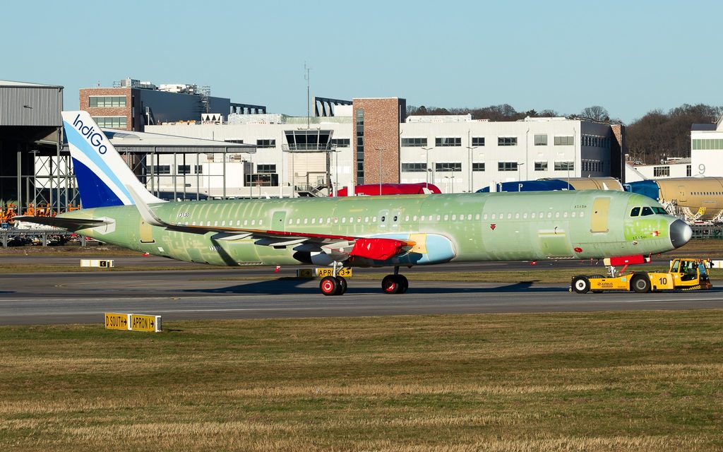 Unfinished Airbus A321neo with an IndiGo tail being towed past factory buildings, with green primer covering its fuselage.