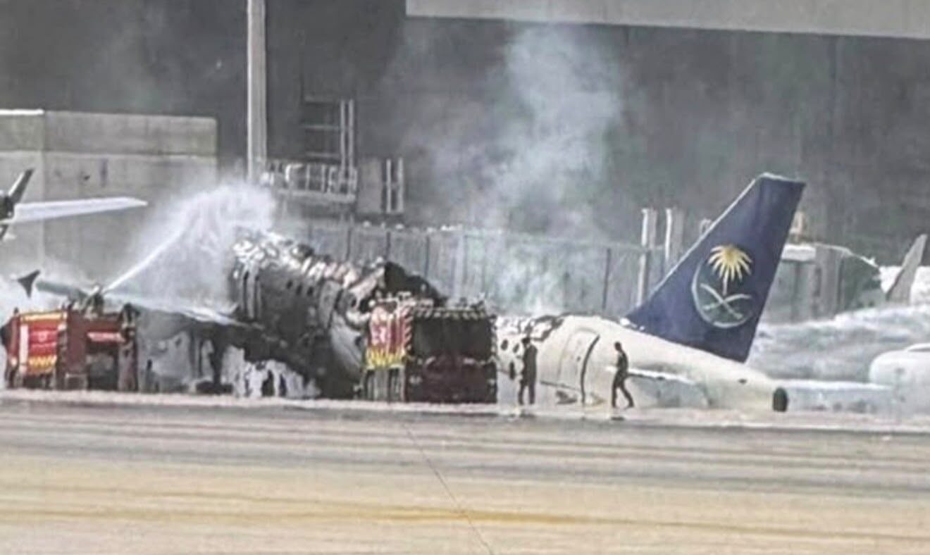 Firefighters spray water onto a severely burnt Saudia aircraft beside an airport terminal, with smoke rising from its exposed fuselage.